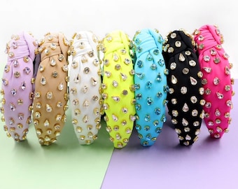 Op de afbeelding: Een verzameling van zeven hoofdbanden in verschillende kleuren, versierd met strass-steentjes en goudkleurige accenten. De kleuren variëren van lila en beige tot wit, neongeel, turquoise, zwart en roze.