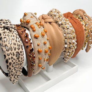 May include: A set of six brown and beige knotted headbands with various embellishments, including rhinestones, pearls, and beads. The headbands are displayed on a white stand.