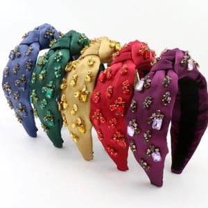 May include: Six jewel-embellished headbands in a row, each a different color: navy blue, emerald green, gold, red, and two shades of purple. The headbands are fabric-covered and adorned with various colored rhinestones.