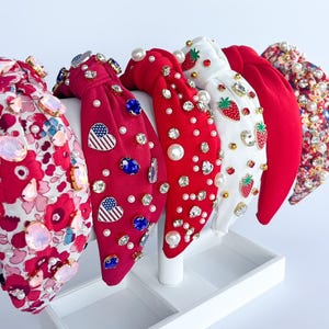 May include: A collection of embellished headbands in various designs. The headbands feature red, white, and floral patterns, adorned with pearls, rhinestones, and other decorative elements. The headbands are displayed on a white stand.