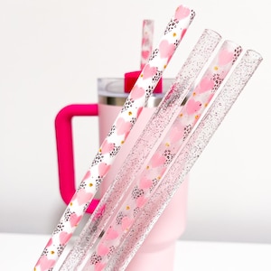 May include: Set of pink and white reusable straws with a heart and leopard print design, and clear glitter straws. The straws are next to a pink tumbler with a stainless steel lid.