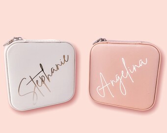 Puede incluir: Dos pequeñas cajas de joyería cuadradas con cierres de cremallera. La caja de la izquierda es blanca con el nombre "Stephanie" escrito en letras doradas. La caja de la derecha es melocotón con el nombre "Angelina" escrito en letras blancas.