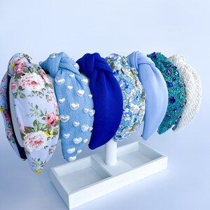 Puede incluir: Exhibici&oacute;n de seis diademas con diversos dise&ntilde;os. Incluyen estampados florales, denim con corazones, azul liso, estampado floral, azul claro y turquesa con lentejuelas. Se presentan en un expositor blanco.