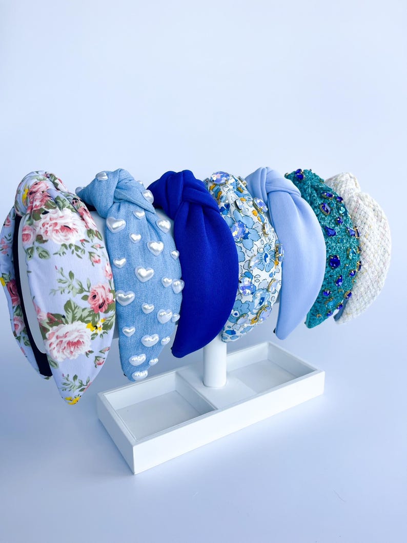 Puede incluir: Exhibici&oacute;n de seis diademas con diversos dise&ntilde;os. Incluyen estampados florales, denim con corazones, azul liso, estampado floral, azul claro y turquesa con lentejuelas. Se presentan en un expositor blanco.