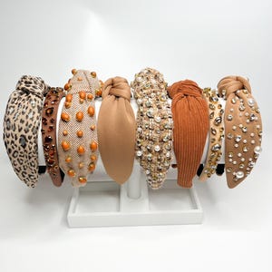 May include: A set of eight brown and beige headbands with various embellishments, including rhinestones, pearls, and fabric textures. The headbands are displayed on a white stand.