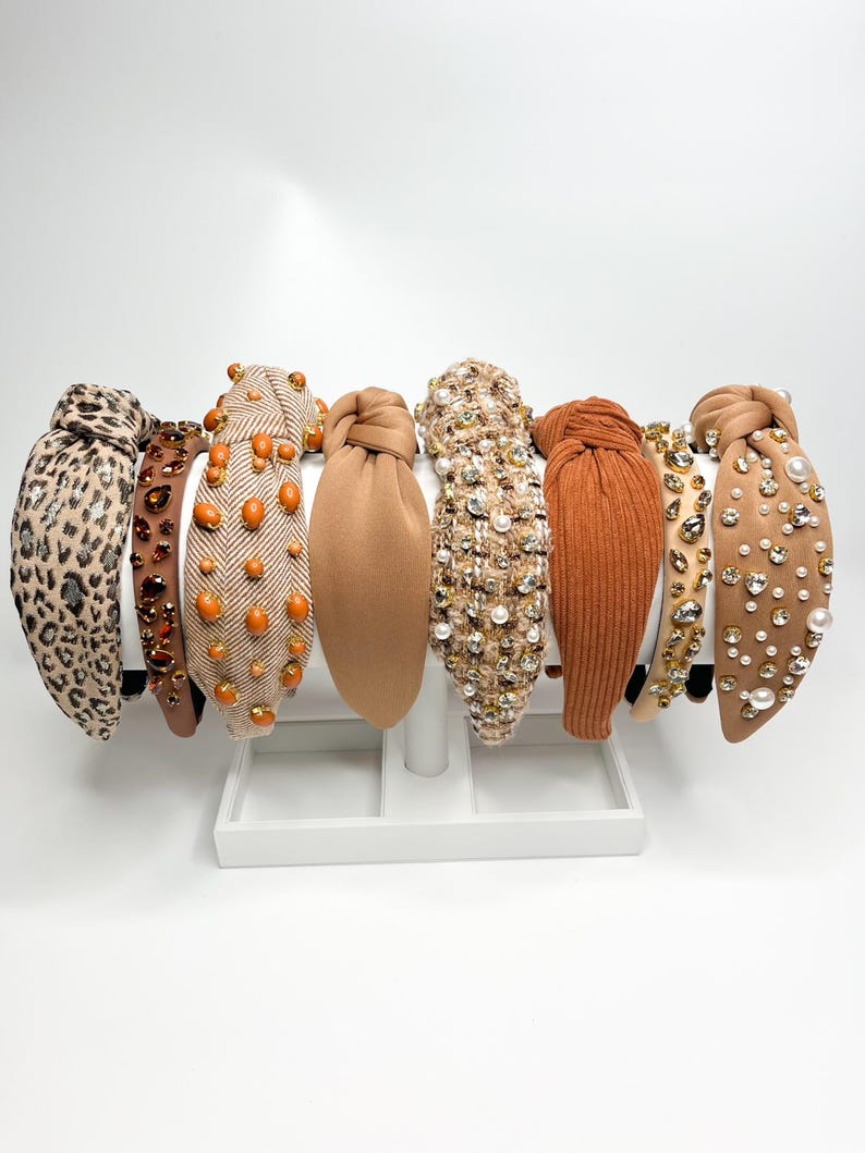 May include: A set of eight brown and beige headbands with various embellishments, including rhinestones, pearls, and fabric textures. The headbands are displayed on a white stand.