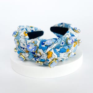 Puede incluir: Una diadema con estampado floral azul claro con grandes adornos de cristal azul claro y transparente. La diadema presenta un dise&ntilde;o anudado y est&aacute; colocada sobre una superficie blanca. Los cristales est&aacute;n enmarcados en oro.