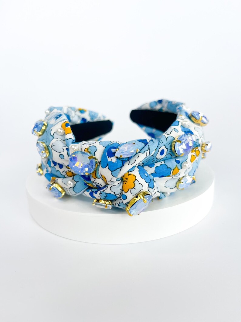 Puede incluir: Una diadema con estampado floral azul claro con grandes adornos de cristal azul claro y transparente. La diadema presenta un dise&ntilde;o anudado y est&aacute; colocada sobre una superficie blanca. Los cristales est&aacute;n enmarcados en oro.