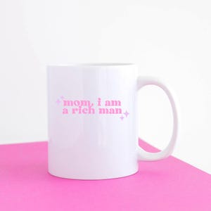 May include: White ceramic mug with a pink design that says "mom, i am a rich man" with pink glitter accents.