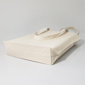 May include: A plain white canvas tote bag with two long straps. The bag is folded and lying on a white surface.