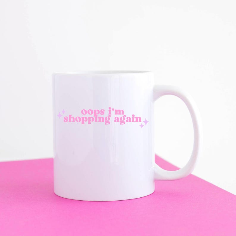 May include: White ceramic mug with a pink inscription that reads "oops i'm shopping again" with pink glitter accents.