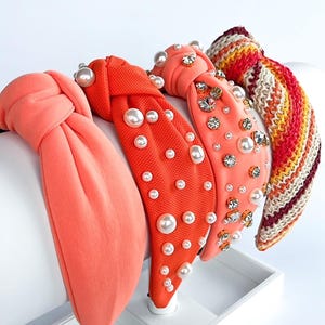 May include: Four headbands in shades of coral and red are displayed. One is solid coral, while the others feature pearl and rhinestone embellishments. The last headband has a striped pattern in red, orange, yellow, and cream.
