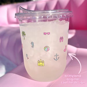 Op de afbeelding: Een doorzichtige plastic beker met deksel gevuld met een lichtgekleurde drank. De beker is voorzien van kleine, kleurrijke iconen met een zomerthema, waaronder een palmboom, hartvormige zonnebrillen, slippers, een zeepaardje en een zandkasteel.