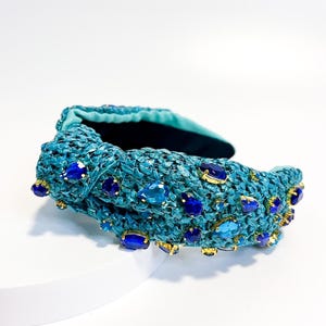 Puede incluir: Una diadema tejida de color turquesa adornada con piedras preciosas azules y zafiro de varios tama&ntilde;os. La diadema presenta un dise&ntilde;o anudado y detalles dorados alrededor de las piedras preciosas. El interior est&aacute; forrado con tela turquesa oscura.
