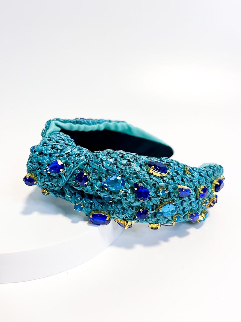 Puede incluir: Una diadema tejida de color turquesa adornada con piedras preciosas azules y zafiro de varios tama&ntilde;os. La diadema presenta un dise&ntilde;o anudado y detalles dorados alrededor de las piedras preciosas. El interior est&aacute; forrado con tela turquesa oscura.