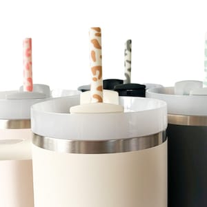 May include: Four stainless steel travel mugs with lids and straws. The mugs are in pink, beige, black, and gray. The straws are patterned with different designs.