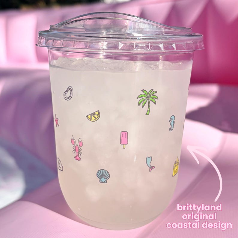 Op de afbeelding: Een doorzichtige plastic beker met deksel gevuld met ijs en een lichtgekleurde drank. De buitenkant is voorzien van kleine, kleurrijke iconen waaronder een palmboom, kreeft, schelp, citroenschijfje, ijsje en zeepaardje. Tekst: brittyland original coastal design.