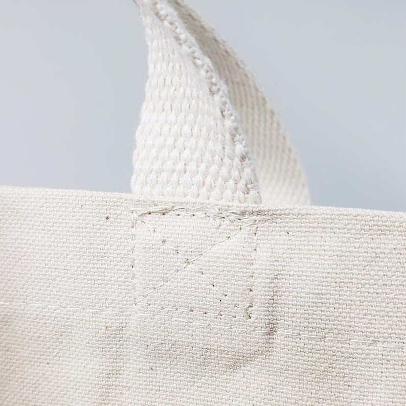 May include: Close-up of a white canvas tote bag with a white cotton webbing handle. The handle is sewn to the bag with white thread.