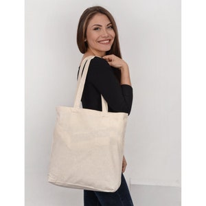 May include: A plain white canvas tote bag with two long handles. The bag is being held by a person wearing a black shirt and blue jeans.