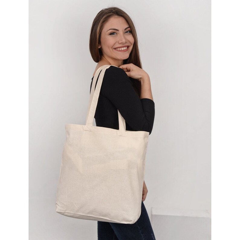 May include: A plain white canvas tote bag with two long handles. The bag is being held by a person wearing a black shirt and blue jeans.