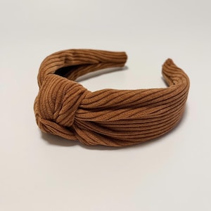May include: A brown corduroy knotted headband. The headband is made of a soft, ribbed fabric and has a knotted design.