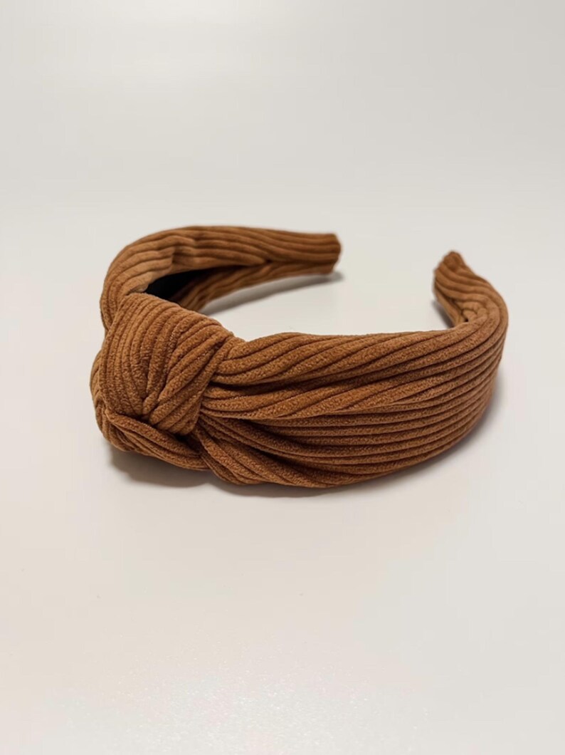 May include: A brown corduroy knotted headband. The headband is made of a soft, ribbed fabric and has a knotted design.