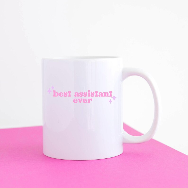 May include: White ceramic mug with a pink inscription that reads "best assistant ever" with pink glitter accents.
