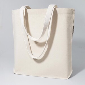 May include: A plain white canvas tote bag with two long straps. The bag has a small label on the side that says "Organic Cotton".
