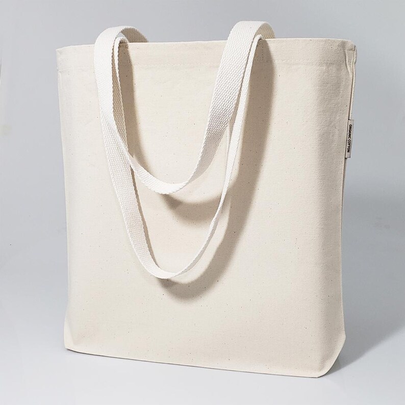 May include: A plain white canvas tote bag with two long straps. The bag has a small label on the side that says "Organic Cotton".