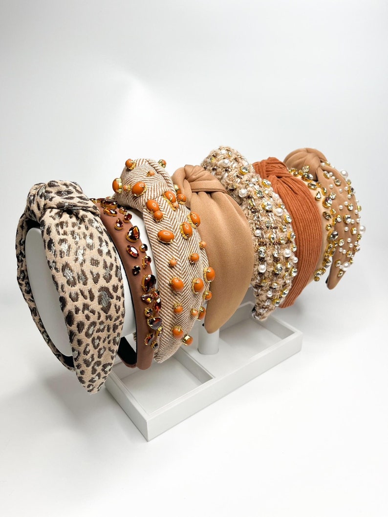May include: A set of six brown and beige knotted headbands with various embellishments, including rhinestones, pearls, and beads. The headbands are displayed on a white stand.