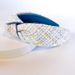 Puede incluir: Una diadema de tweed blanco con un nudo en la parte superior. La diadema presenta un patr&oacute;n tejido con motas amarillas, rosas y azules. El interior est&aacute; forrado con una tela azul vibrante. Un elegante accesorio para el cabello.
