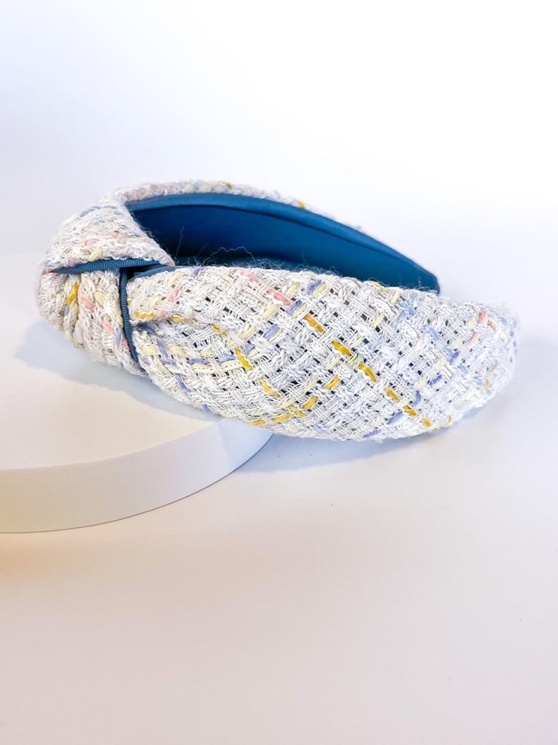 Puede incluir: Una diadema de tweed blanco con un nudo en la parte superior. La diadema presenta un patr&oacute;n tejido con motas amarillas, rosas y azules. El interior est&aacute; forrado con una tela azul vibrante. Un elegante accesorio para el cabello.