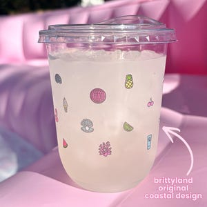Op de afbeelding: Een doorzichtige plastic koude beker met deksel, gevuld met ijs en een lichtgekleurde drank. De buitenkant is voorzien van kleine kleurrijke iconen zoals een discobal, ananas, schelp, koraal en limoenschijfje. Tekst: brittyland original coastal design.
