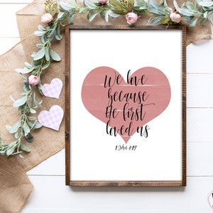 May include: A printable wall art design with a pink heart and the text "We love because He first loved us 1 John 4:19".