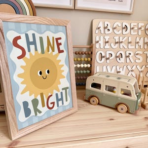 Puede incluir: Una impresi&oacute;n enmarcada con las palabras "SHINE BRIGHT" y una ilustraci&oacute;n de un sol sonriente. La impresi&oacute;n est&aacute; en un marco de madera clara y se encuentra sobre una superficie de madera. Otros art&iacute;culos incluyen una furgoneta camper de juguete de madera y un rompecabezas de alfabeto.