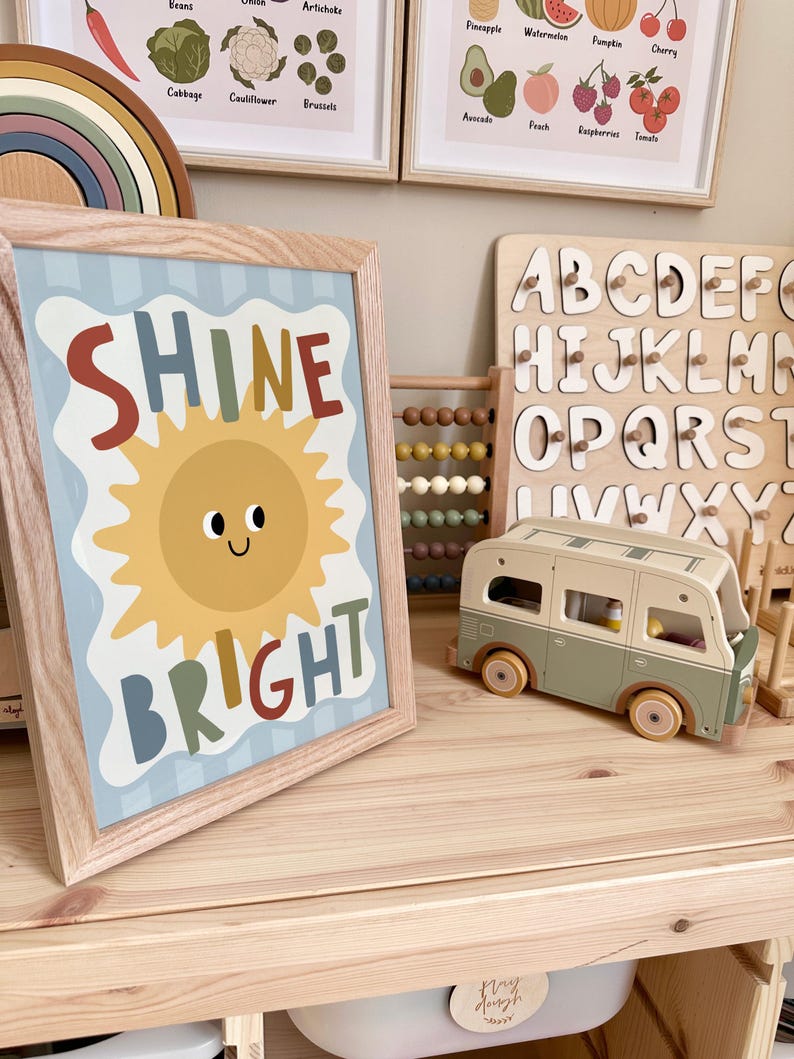 Puede incluir: Una impresi&oacute;n enmarcada con las palabras "SHINE BRIGHT" y una ilustraci&oacute;n de un sol sonriente. La impresi&oacute;n est&aacute; en un marco de madera clara y se encuentra sobre una superficie de madera. Otros art&iacute;culos incluyen una furgoneta camper de juguete de madera y un rompecabezas de alfabeto.