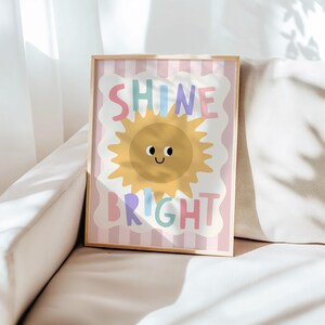 Puede incluir: Un estampado enmarcado con las palabras "SHINE BRIGHT" en letras coloridas. La obra de arte presenta un sol amarillo sonriente con un borde festoneado blanco sobre un fondo a rayas rosas y blancas. El marco es de color madera clara.