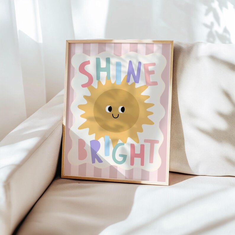 Puede incluir: Un estampado enmarcado con las palabras "SHINE BRIGHT" en letras coloridas. La obra de arte presenta un sol amarillo sonriente con un borde festoneado blanco sobre un fondo a rayas rosas y blancas. El marco es de color madera clara.
