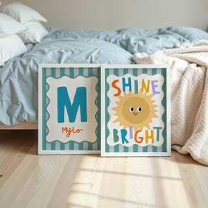 Puede incluir: Dos impresiones art&iacute;sticas enmarcadas. Una presenta la letra "M" turquesa con el nombre "Mylo" en naranja. La otra dice "Shine Bright" con una ilustraci&oacute;n de sol sonriente. Ambas tienen marcos blancos y fondos a rayas turquesas.