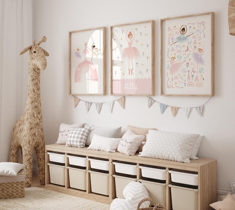 May include: A light wood bench with storage bins and a light gray cushion. Three framed prints with illustrations of ballerinas in pink dresses and floral designs hang on the wall above the bench. A light brown giraffe figurine stands to the left of the bench.