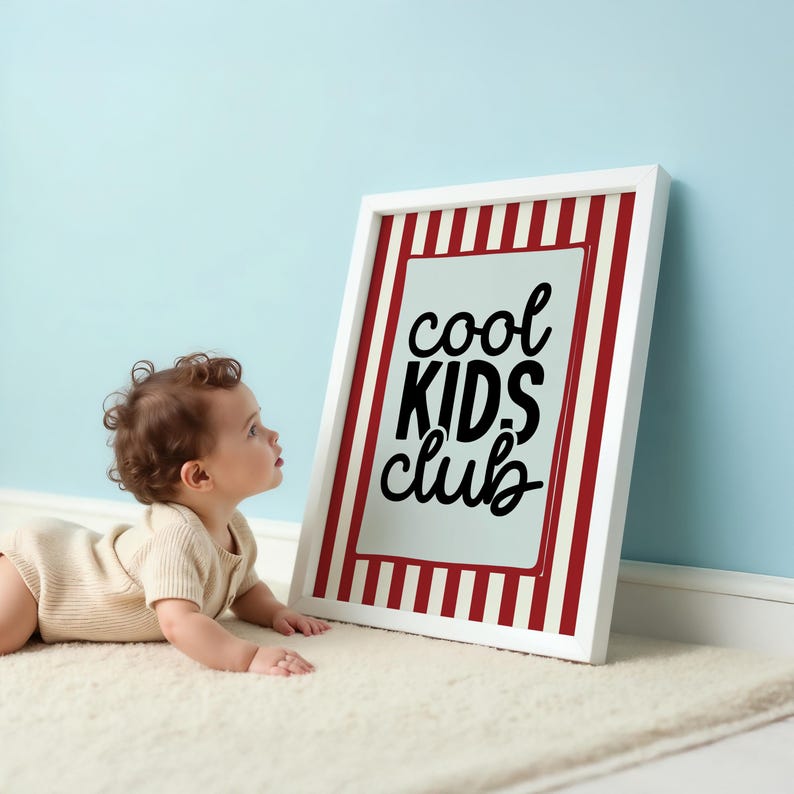 May include: A white framed sign with red and white stripes and the words "cool KIDS club" in black. A baby in a beige onesie is lying on a white rug, looking at the sign. The background is a light blue wall.
