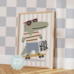 May include: A framed print featuring a cartoon alligator wearing sunglasses, a striped shirt, and blue shorts, riding a skateboard. The text "IT'S A WILD RIDE" is displayed on the right side of the image. The print is in a wooden frame.