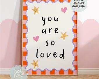 May include: A framed art print with the words "you are so loved" in black script. The print has a white background with pink hearts and yellow star illustrations. The frame has a pink and orange checkered border.