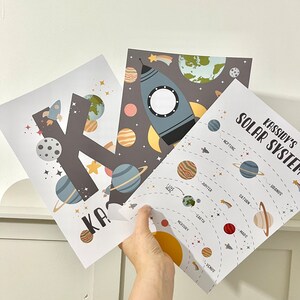 Pode incluir: Dois cartazes imprim&iacute;veis com tema espacial. Um cartaz apresenta a letra K com planetas e estrelas. O outro cartaz mostra um foguete e um diagrama do sistema solar com os nomes dos planetas: Merc&uacute;rio, V&eacute;nus, Terra, Marte, J&uacute;piter, Saturno, Urano e Neptuno. O texto "Kassidy's Solar System" est&aacute; no topo do cartaz.