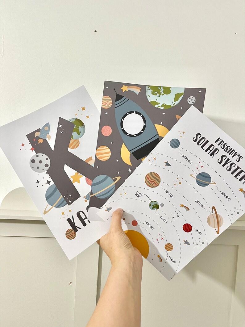 Pode incluir: Dois cartazes imprim&iacute;veis com tema espacial. Um cartaz apresenta a letra K com planetas e estrelas. O outro cartaz mostra um foguete e um diagrama do sistema solar com os nomes dos planetas: Merc&uacute;rio, V&eacute;nus, Terra, Marte, J&uacute;piter, Saturno, Urano e Neptuno. O texto "Kassidy's Solar System" est&aacute; no topo do cartaz.