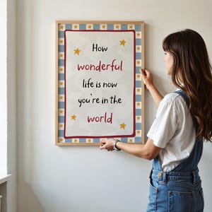 May include: A framed print with the words "How wonderful life is now you're in the world" in red and black text. The print has a white background with yellow stars and a blue and white checkered border. The frame is a light brown color.