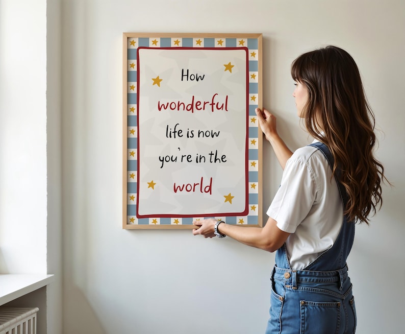 May include: A framed print with the words "How wonderful life is now you're in the world" in red and black text. The print has a white background with yellow stars and a blue and white checkered border. The frame is a light brown color.