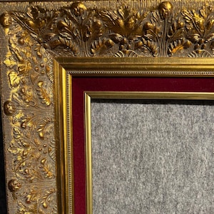 May include: Ornate gold picture frame with a burgundy mat and a gray backing. The frame features detailed floral and leaf carvings. The inner frame is a rich gold color, and the mat is a deep burgundy.