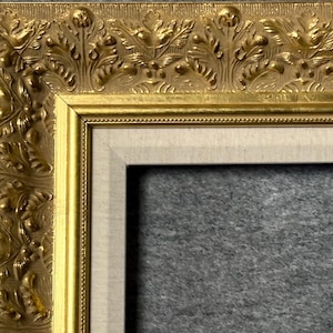 May include: A gold picture frame with an ornate, embossed design featuring leaves and flowers. The frame has a white mat and a gray background.