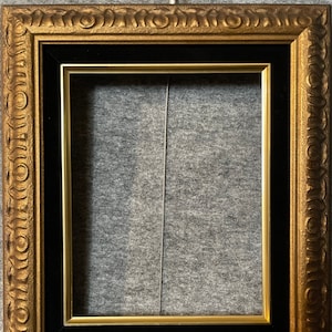 May include: A gold picture frame with a black mat and ornate detailing around the edges.